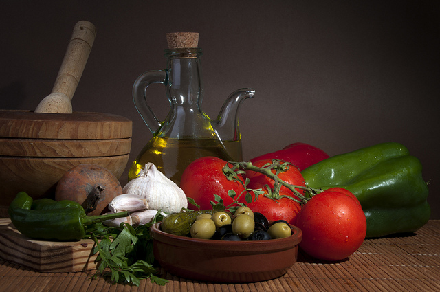 Fresh vegetables including tomatoes, garlic, green peppers, and herbs, alongside a bottle of olive oil and a bowl of olives, arranged on a wooden surface, emphasizing healthy ingredients for meal preparation.