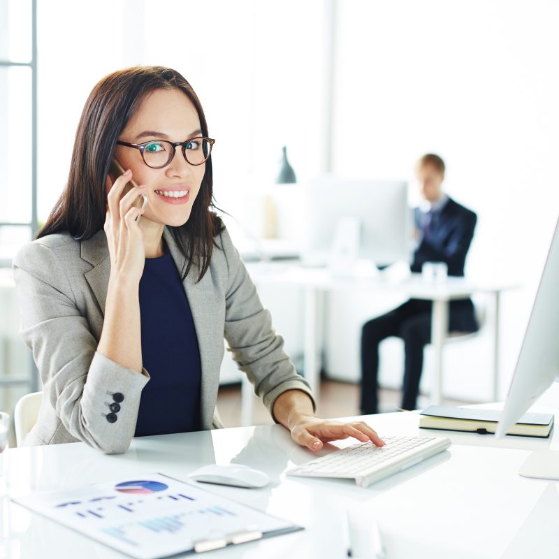 Professional woman on phone in office, smiling while using computer and keyboard, with charts and documents on desk, emphasizing workplace productivity and health insurance discussions.