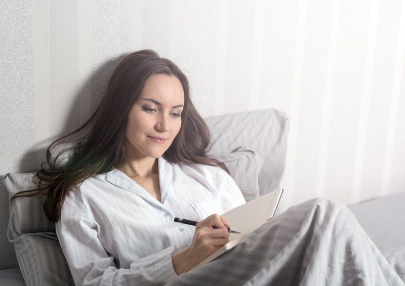 Woman in pajamas writing in a notebook while relaxing in bed, representing wellness and self-care in health-related content.