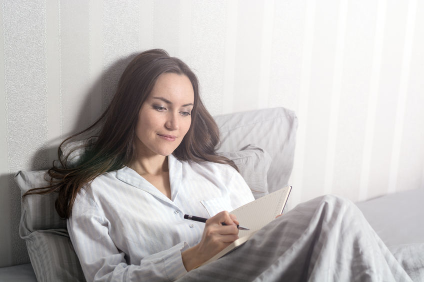 Woman in pajamas writing in a notebook while sitting in bed, emphasizing morning routine and mindfulness for well-being.