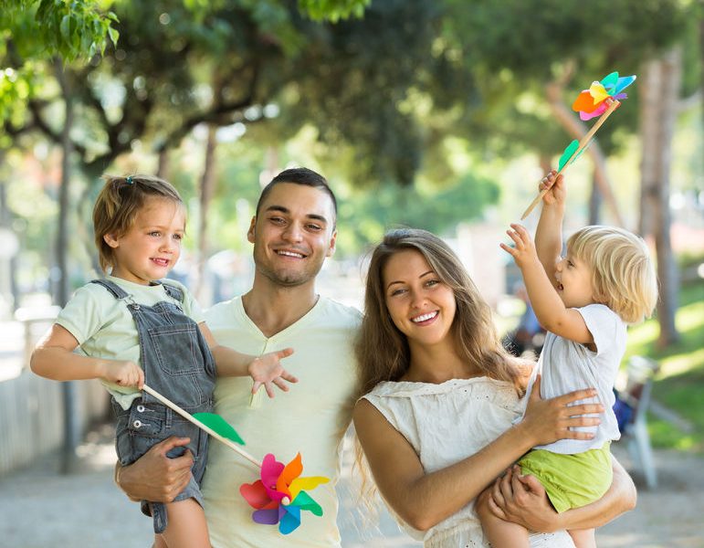 Family enjoying a day outdoors, with parents holding children and colorful pinwheels, reflecting themes of family wellness and happiness in the context of health insurance discussions.
