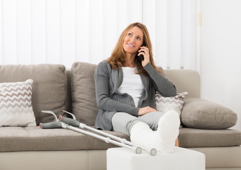 Woman on couch with crutches and foot in a cast, talking on the phone in a cozy living room setting, illustrating recovery and wellness.