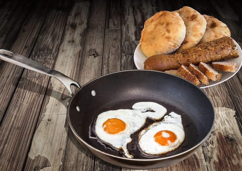 Fried eggs with bright yolks in a frying pan, accompanied by a plate of assorted breads, including pita and sliced whole grain, on a wooden countertop.