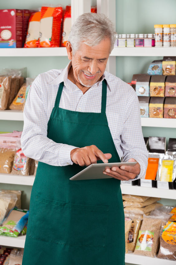 Older man in a green apron using a tablet in a grocery store, surrounded by various packaged food items, illustrating digital grocery planning for healthier eating habits.