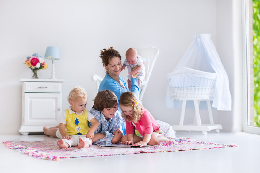 Family enjoying time in a bright nursery, mother holding a baby, children playing on a colorful rug, with a crib and decorative elements in the background.