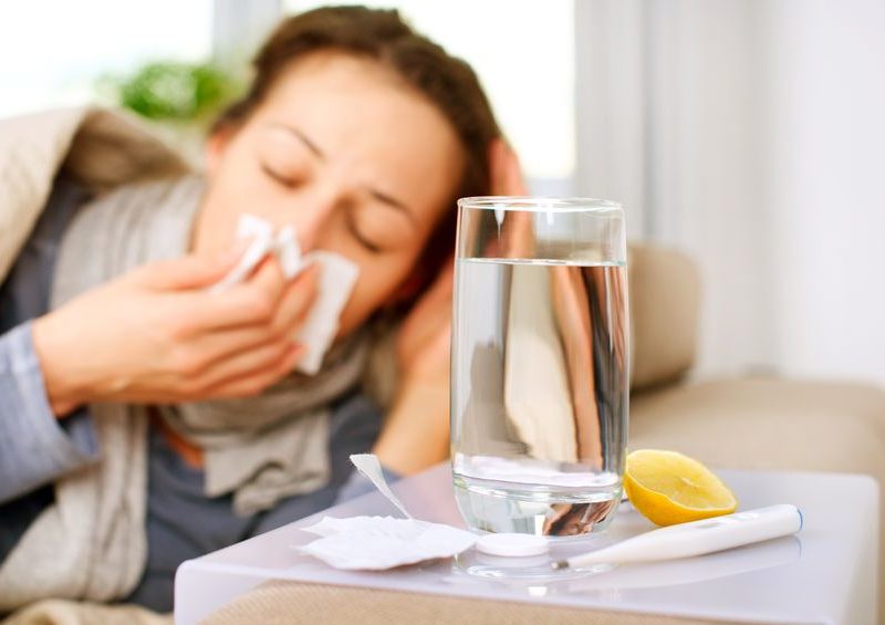 Woman with a cold holding tissue, glass of water with lemon, and thermometer on table, depicting tips for surviving cold and flu season.