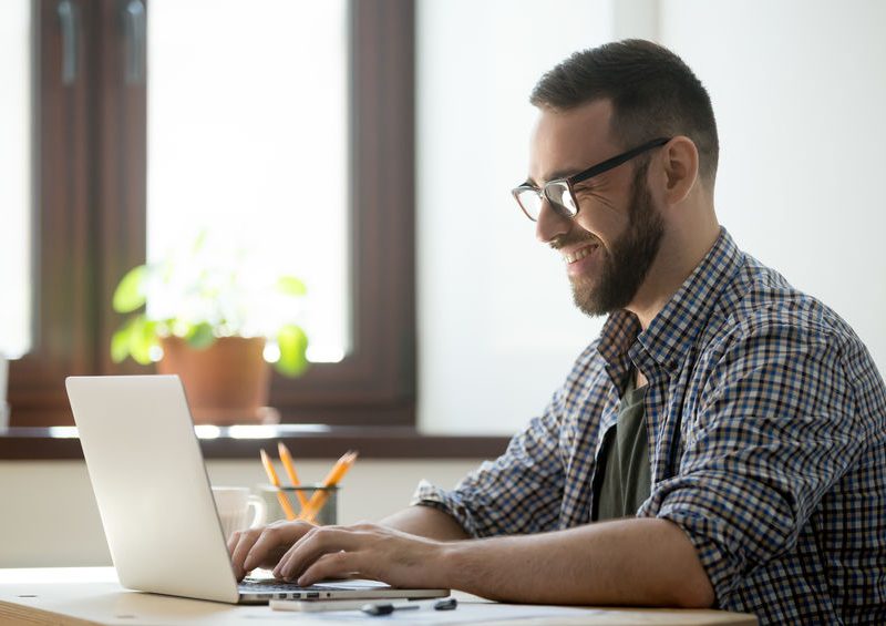 Man smiling while working on a laptop at home, surrounded by a plant and stationery, illustrating productivity and health-focused living.