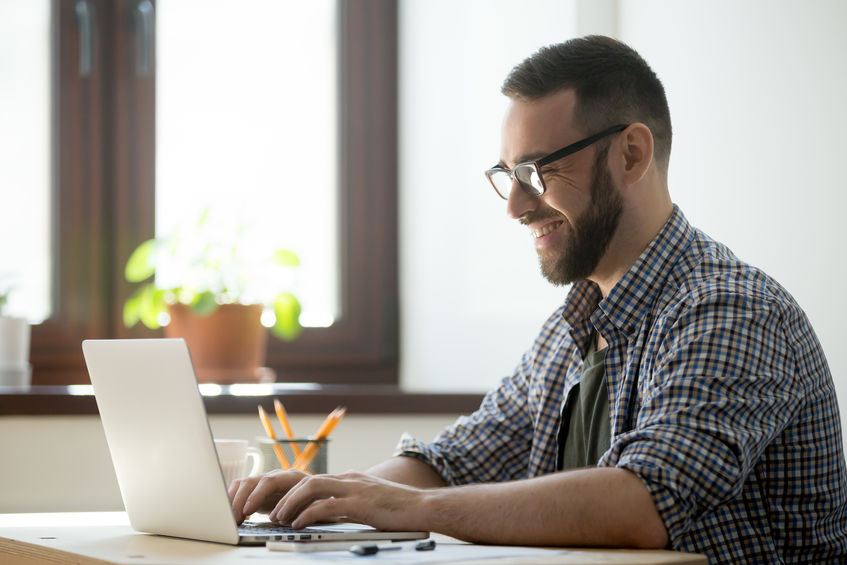 Man smiling while using a laptop in a bright, modern workspace, emphasizing productivity and focus on health-related topics.