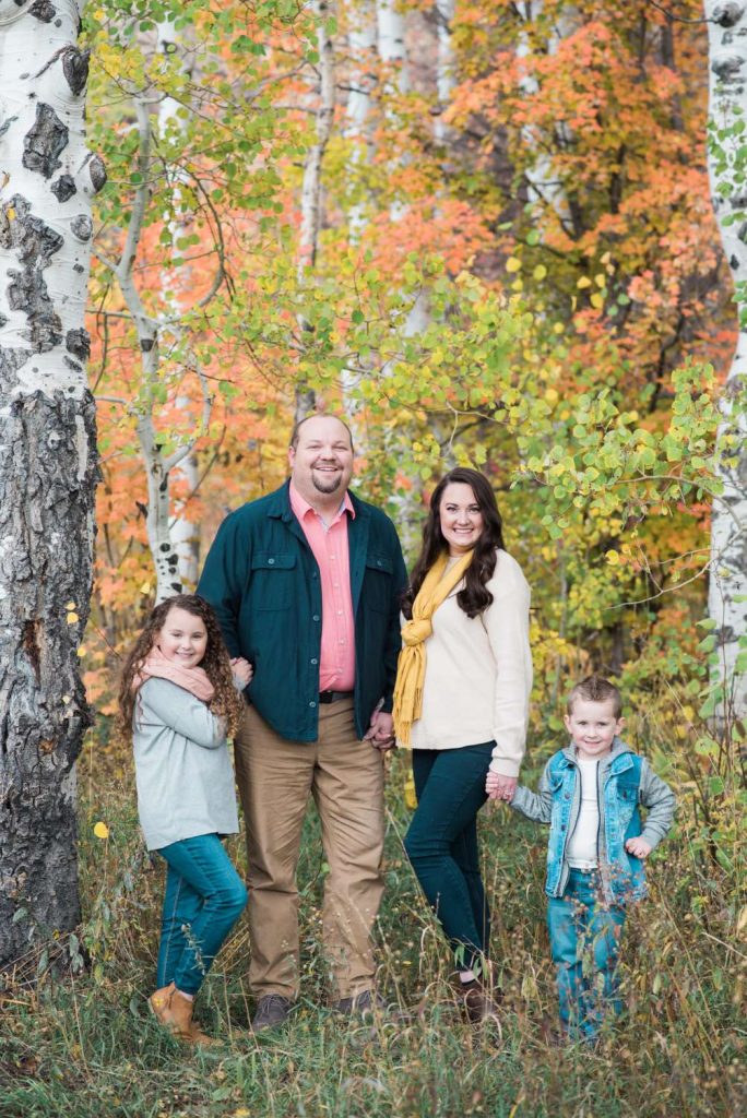 Family portrait in autumn setting, featuring a woman in a cream sweater and yellow scarf, a man in a green jacket, and two children, one girl and one boy, surrounded by colorful fall foliage, representing a warm family atmosphere.