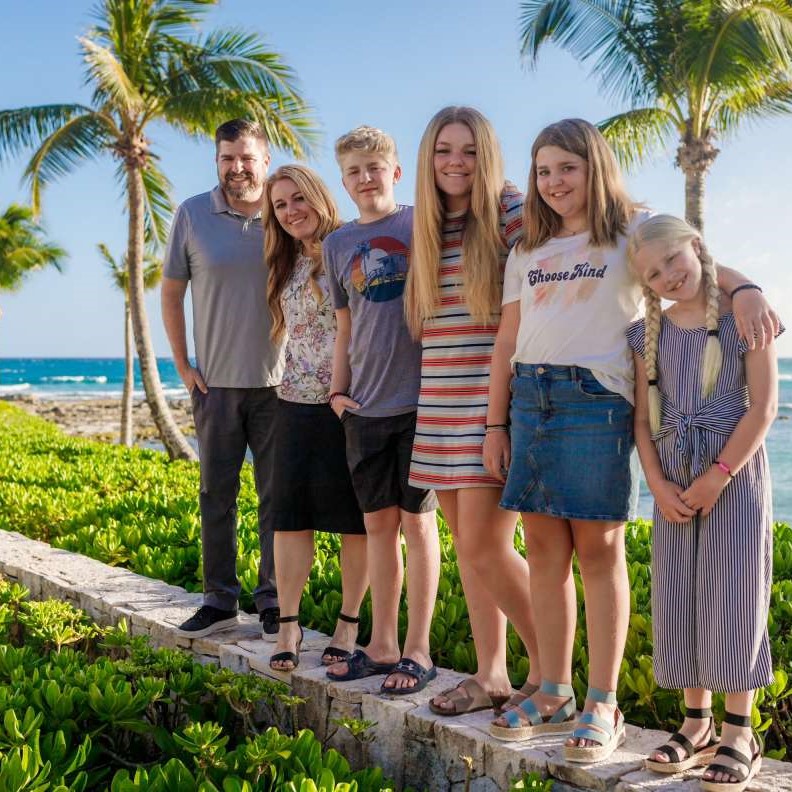 Family posing together outdoors near the ocean, surrounded by palm trees and greenery, showcasing a friendly and relaxed atmosphere.