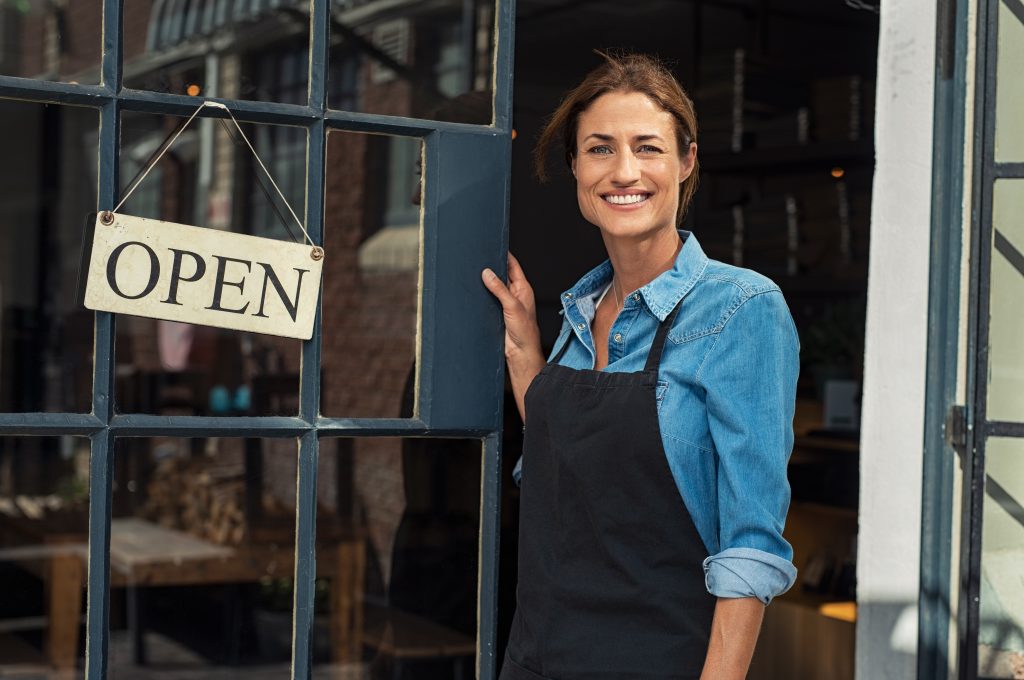 Smiling woman in apron standing at an open shop door, representing entrepreneurship and accessible healthcare support for self-employed individuals seeking health insurance solutions.