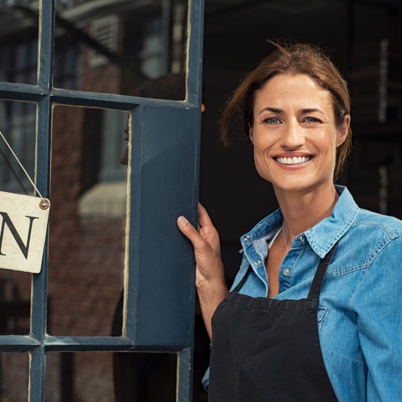 Smiling business owner in denim shirt and apron standing at the entrance of a shop with an "OPEN" sign, representing entrepreneurship and small business success.