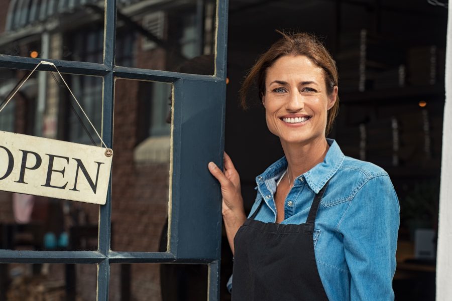 Smiling business owner standing at the entrance of a shop, holding an open sign, representing entrepreneurship and health insurance guidance.