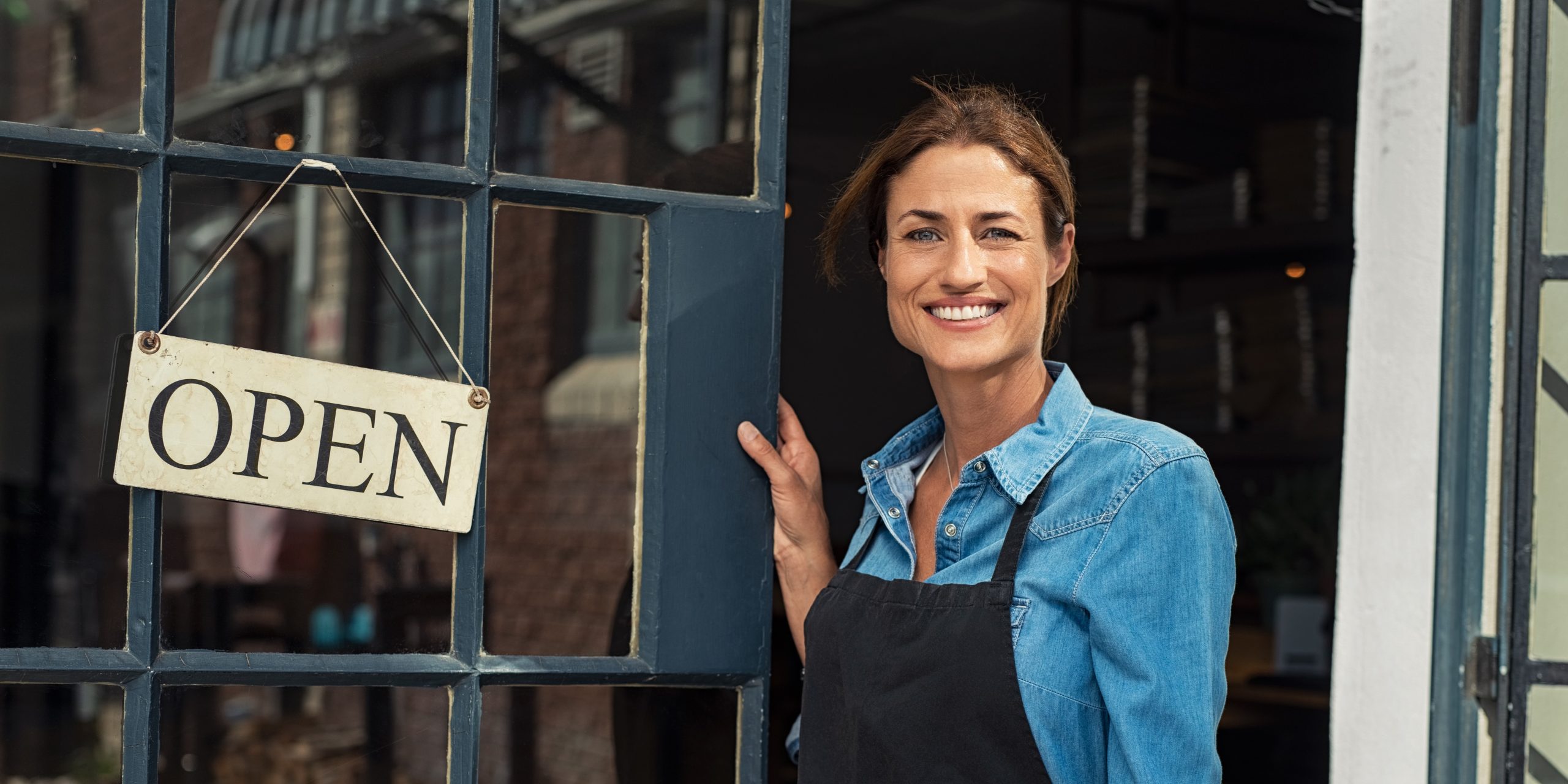 Smiling woman standing at the entrance of a business, holding an "OPEN" sign, representing self-employed individuals seeking health insurance options.