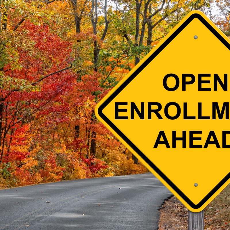 Open enrollment ahead sign on a winding road surrounded by vibrant autumn foliage, representing the upcoming health insurance enrollment period.