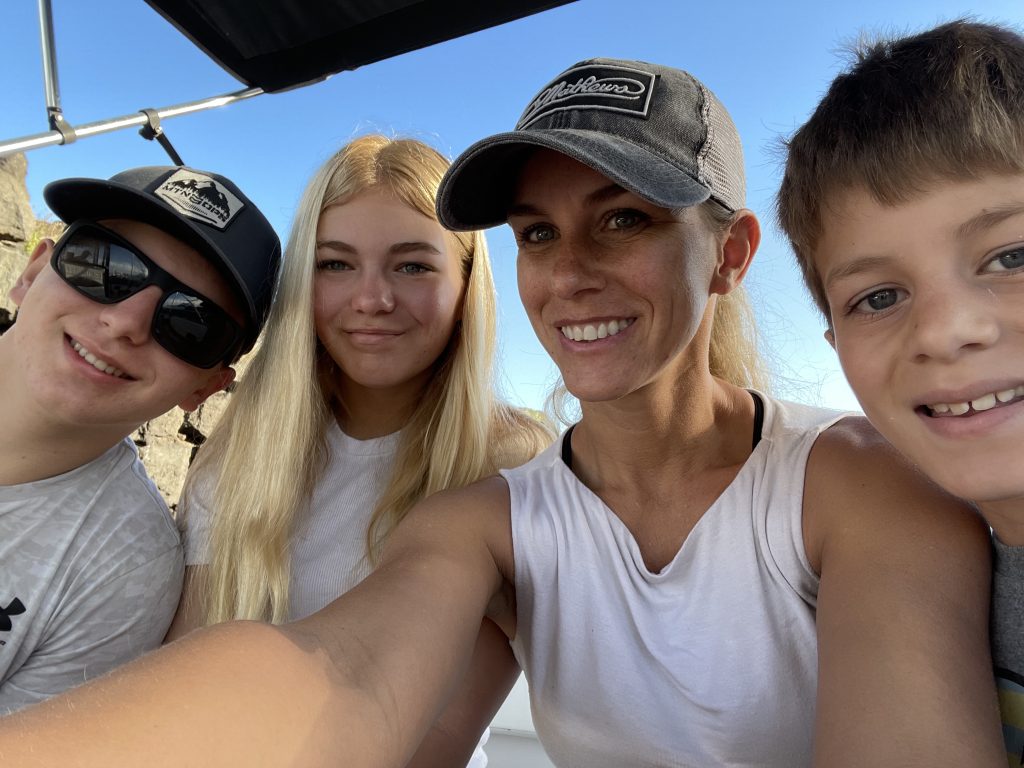 Group selfie of a woman and three children outdoors, smiling together, with blue sky and boat setting, reflecting a family-oriented atmosphere.