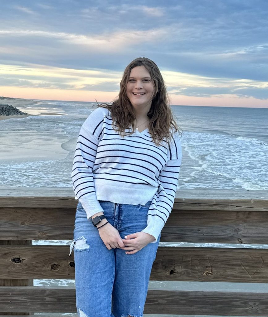 Young woman smiling on a wooden pier by the beach, wearing a striped sweater and jeans, with ocean waves and a colorful sky in the background.